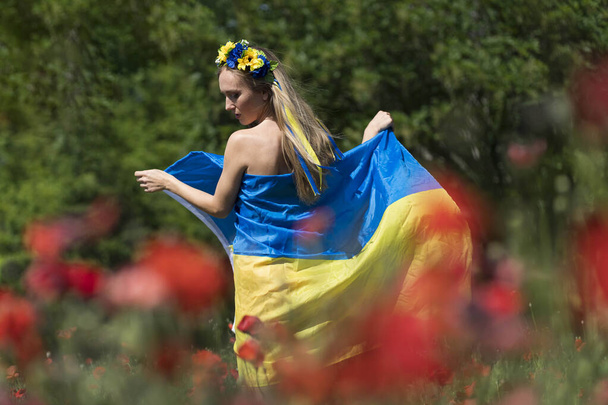 Una joven rubia ucraniana se encuentra en un campo de flores de amapola roja sosteniendo la bandera de Ucrania mostrando su apoyo a la guerra en su país natal, Ucrania. - Foto, Imagen
