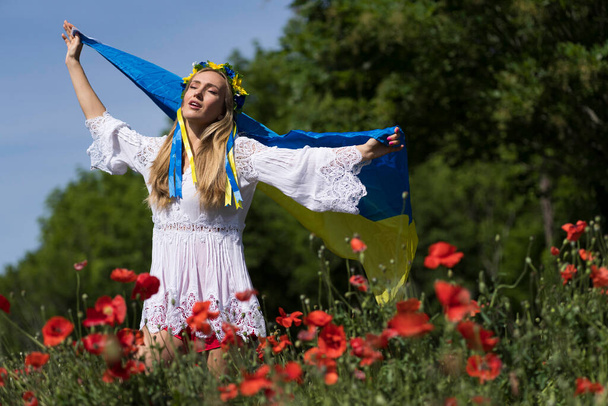 Eine junge blonde Ukrainerin steht auf einem Feld mit roten Mohnblumen und hält die Fahne der Ukraine in der Hand, die ihre Unterstützung für den Krieg in ihrem Heimatland Ukraine zeigt. - Foto, Bild
