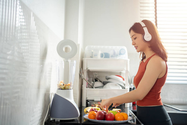 The sportswoman making smoothies from fruits in the kitchen at home while listening to music through headphones - lifestyles concepts - 写真・画像