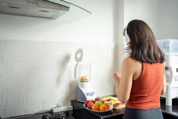 The sportswoman making smoothies from fruits in the kitchen at home while listening to music through headphones - lifestyles concepts - 写真・画像