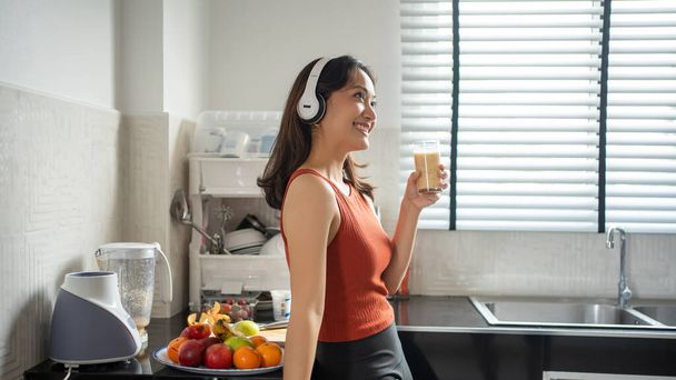 Beautiful young woman making and drinking smoothies from fruits in the kitchen at home while listening to music through headphones - lifestyles concepts - Фото, изображение