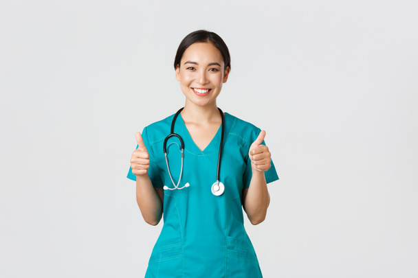 Covid-19, healthcare workers, pandemic concept. Professional confident smiling doctor, female physician in scrubs showing thumbs-up with assured expression, ensure all good, perfect or excellent - Photo, Image