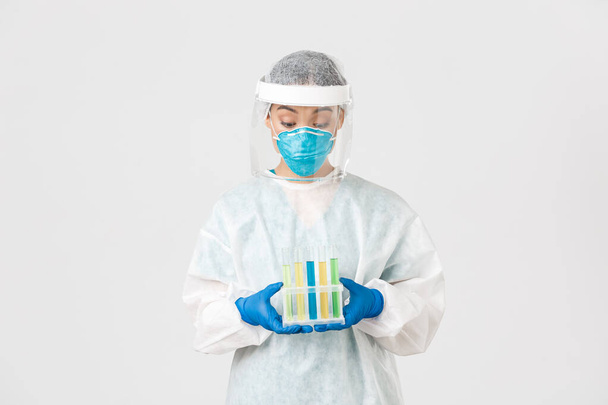 Covid-19, coronavirus disease, healthcare workers concept. Excited asian female doctor, tech lab in personal protective equipment holding test-tubes with vaccine or analyses, white background - Photo, Image