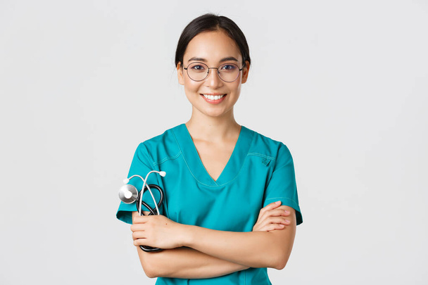 Covid-19, doença coronavírus, conceito de trabalhadores de saúde. Close-up de profissional confiante médico feminino, enfermeira de óculos e esfrega de pé fundo branco, braços cruzados
 - Foto, Imagem