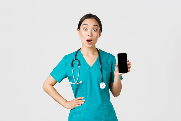 Covid-19, zdravotničtí pracovníci a koncept on-line medicíny. vzrušený a ohromený asijské ženy zdravotní sestra, lékař vypadat překvapeně při zobrazování obrazovky mobilního telefonu, internetové konzultace app, bílé pozadí - Fotografie, Obrázek