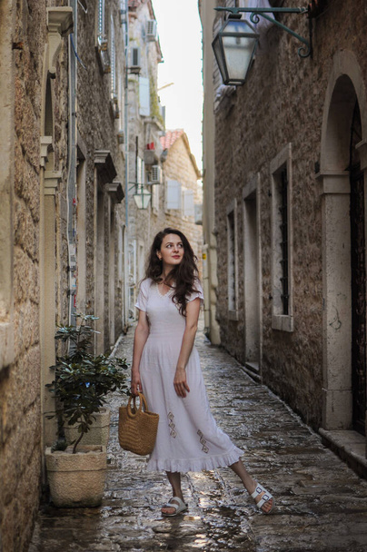 A young beautiful woman in a dress is walking in a tiny street with stone buildings in the old town of Budva, Montenegro - Fotó, kép
