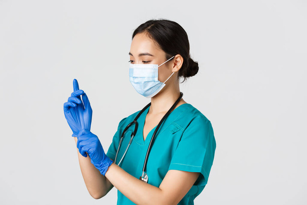 Covid-19, coronavirus disease, healthcare workers concept. Professional smiling asian female nurse, physician in scrubs and medical mask put on rubber gloves for checkup, patient examination - Photo, image