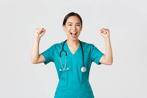 Covid-19, healthcare workers and preventing virus concept. Successful confident asian female doctor or nurse in scrubs fist pump and shouting yes, rejoicing, encourage herself, aim for victory - Photo, Image