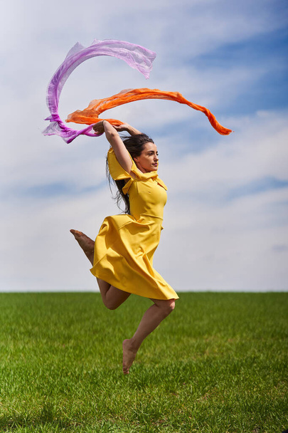 Action shot of a young attractive woman in yellow dress jumping for joy on a green wheat field in a sunny day - Fotoğraf, Görsel