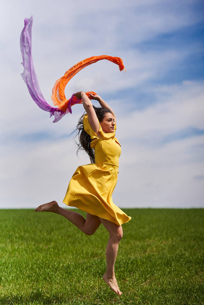 Action shot of a young attractive woman in yellow dress jumping for joy on a green wheat field in a sunny day - Foto, afbeelding