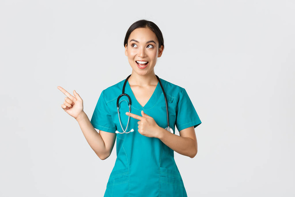 Covid-19, Gesundheitspersonal, Pandemiekonzept. Optimistisch lächelnde asiatische Krankenschwester, Ärztin im Peeling, schaut und zeigt in die linke obere Ecke, zeigt Banner, demonstriert Ansage - Foto, Bild