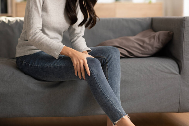 A woman is sitting on the grey couch and try to do self massage on her thigh and joint as well. She squeezes the muscle to relief her painful. Massage could help her to get better from ache.Knee pain - Photo, Image