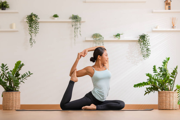 Side view Asian woman wearing green sportwear doing Yoga exercise.Yoga One Legged King Pigeon pose or Eka Pada Rajakapotasana.Calm of healthy young female breathing and meditation yoga at cozy home - 写真・画像