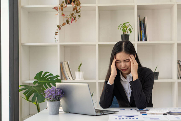 Asian businesswoman having a headache during work analyzing financial graphs with laptop at office desk - Foto, afbeelding