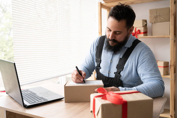 Indian male business owner working from home online shopping Concept of entrepreneur and online trading. Sitting at home in the workplace with a laptop in work overalls and writes the delivery address - Foto, Imagen