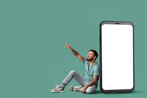 Cool tattooed young man sitting near big smartphone and pointing at something on green background - Photo, Image