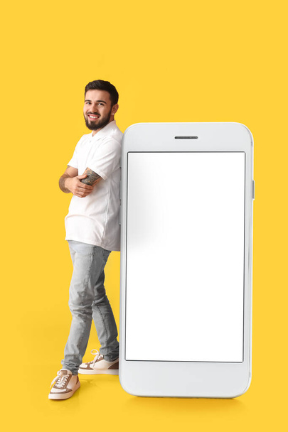 Cool tattooed young man near big smartphone on yellow background - Photo, Image