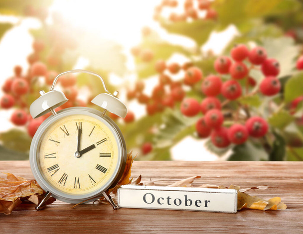 Reloj despertador, palabra OCTUBRE y hojas de otoño en la mesa al aire libre. Fin del horario de verano - Foto, imagen