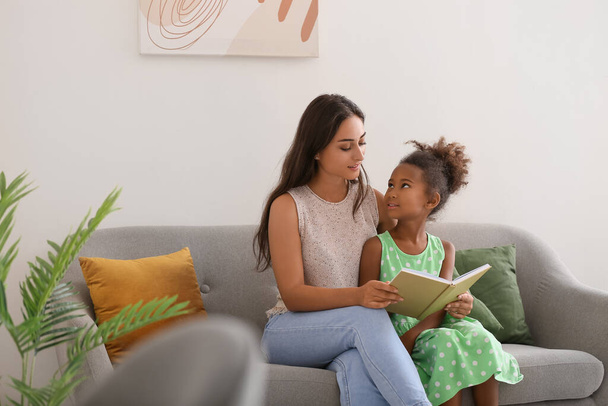 Little African-American girl reading book with her teacher on sofa - Photo, image