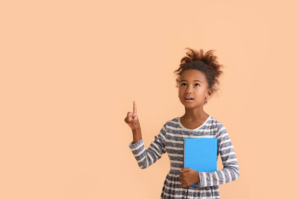 Little African-American girl with book pointing at something on beige background - Fotoğraf, Görsel