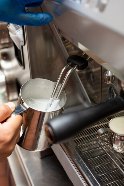 Uitzicht op een hand met een roestvrijstalen kruik vol gestoomde melk, naast een espressomachine, in een caféomgeving. - Foto, afbeelding