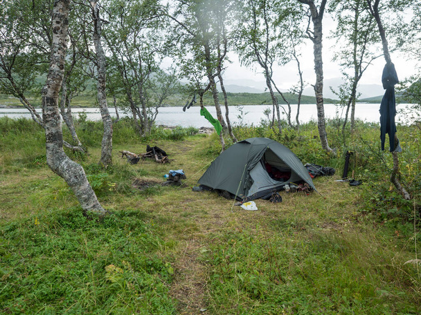 Small open green tent with backpack and hiking gear on grassy shore of Tarra river with birch trees. Swedish Lapland Landscape with green hills at Padjelantaleden hiking trail. Summer day - Foto, immagini