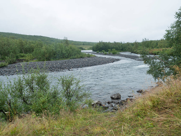 View of winding Tarra river with birch bush, gravel, grass and granite rock. Northern landscape in Swedish Lapland at Padjelantaleden hiking trail. Moody, rainy day, cloudy sky, summer. - Photo, Image