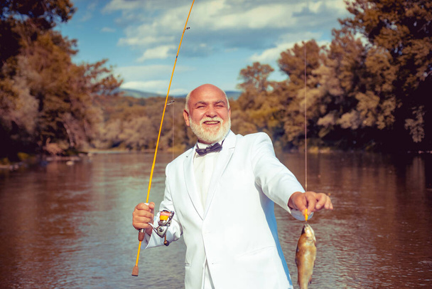 Fisherman caught a trout fish. Portrait of cheerful smiling angler senior man fishing. Grandfather with catch fish. Mature man fisherman in white suit and bowtie with fishing rod. - Foto, immagini