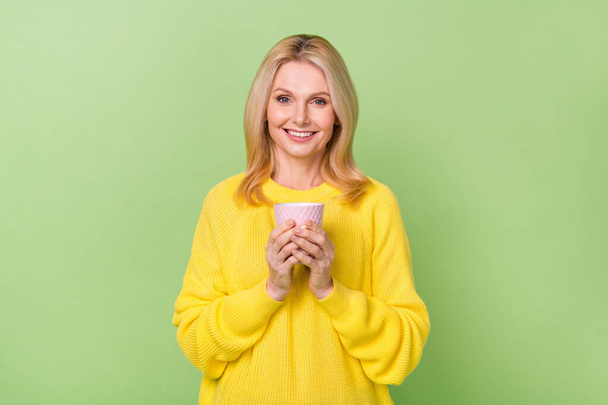Photo of positive aged lady hold latte enjoying refreshment wear yellow sweater isolated green color background - Fotoğraf, Görsel