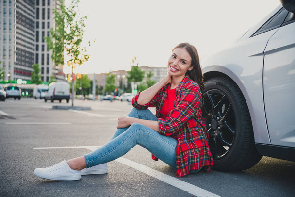 Portrait of attractive cheerful girl driver sitting on floor near new modern car ownership dealer on parking outdoors - Fotografie, Obrázek