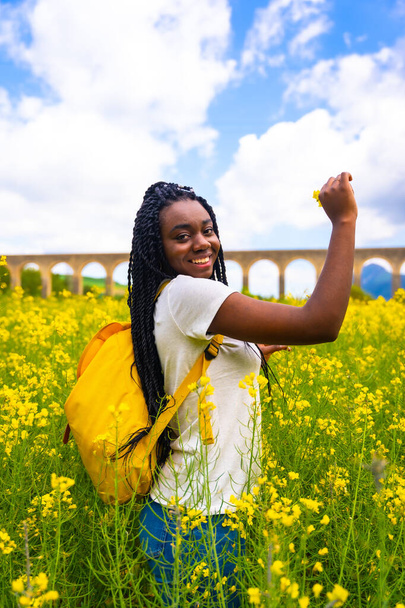 Breathing fresh air and the scent of the flower, a black ethnic girl with braids, a traveler, in a field of yellow flowers - Foto, Imagem