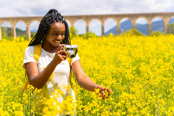 Taking photos with a vintage camera, a black ethnic girl with braids, a traveler, in a field of yellow flowers - Foto, imagen