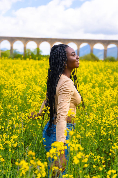 Lifestyle, enjoying nature smiling, portrait of black ethnic girl with braids, traveler, in a field of yellow flowers - Photo, Image