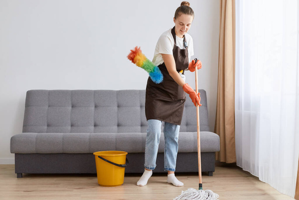 Portrait of young adult woman wearing jeans, white t shirt and apron washing floor with mop, standing in living room near sofa, cleaning her flat, doing household chores. - Photo, Image