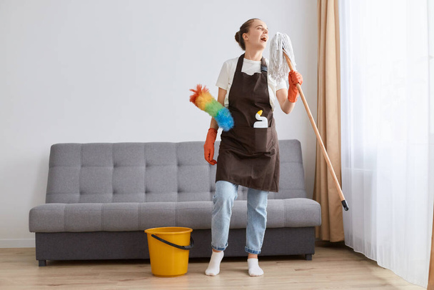 Indoor shot of happy positive housewife woman cleaning floor with mop at home, singing and having fun while cleaning her house, standing near sofa and yellow bucket. - Zdjęcie, obraz