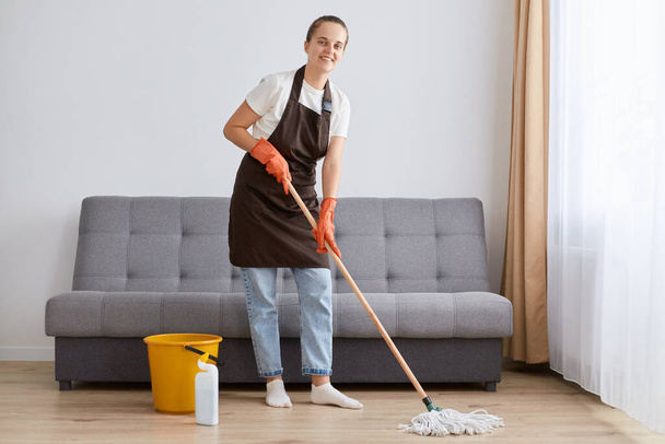 Horizontal shot of attractive Caucasian woman wearing casual clothing and brown apron washing floor, holding mop and looking at camera, cleaning living room. - Photo, image