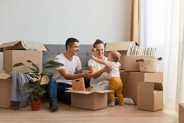 Indoor shot of happy family unpacking moving box at their new house, sitting on floor surrounded with cardboard boxes, playing with infant baby while moving in new apartment. - Foto, Imagen