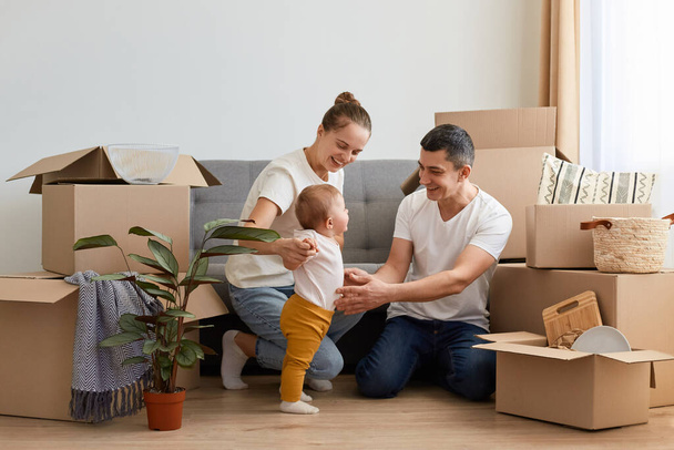 Image of positive family having rest while unpacking moving box at their new house, sitting on floor near sofa and playing with their infant kid, smiling happily. - Fotografie, Obrázek