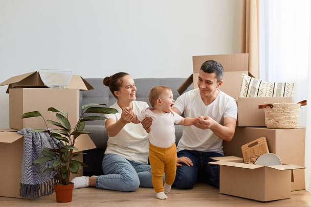 Portrait of happy loving couple with their baby kid moving in new flat, sitting on floor near cough and having fun with their infant daughter, posing among cardboard boxes with belongings. - Φωτογραφία, εικόνα
