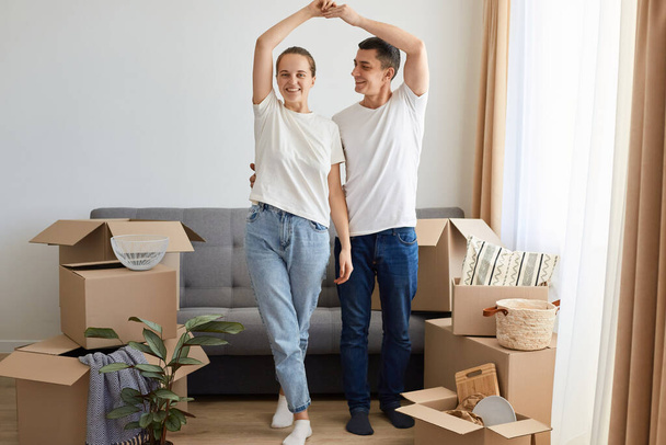 Full length portrait of woman and man wearing jeans and white t shirts posing among carton parcels with belongings, standing near sofa and making roof with hands, moving in a new flat. - Foto, Imagem