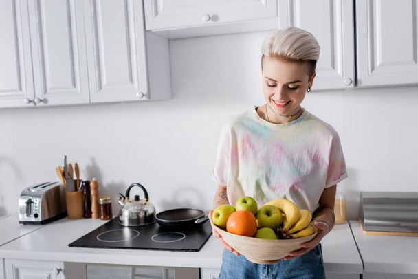 cheerful woman with trendy hairstyle holding bowl of ripe fruits in kitchen - Foto, Imagem
