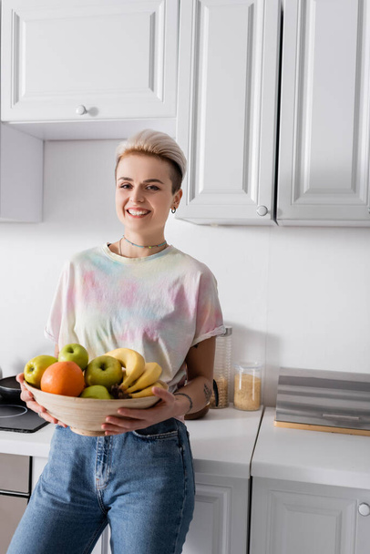 cheerful and stylish woman with bowl of ripe fruits smiling at camera in kitchen - Fotó, kép