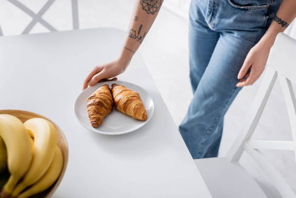 partial view of tattooed woman in jeans near tasty croissants and blurred bananas - Foto, Imagem