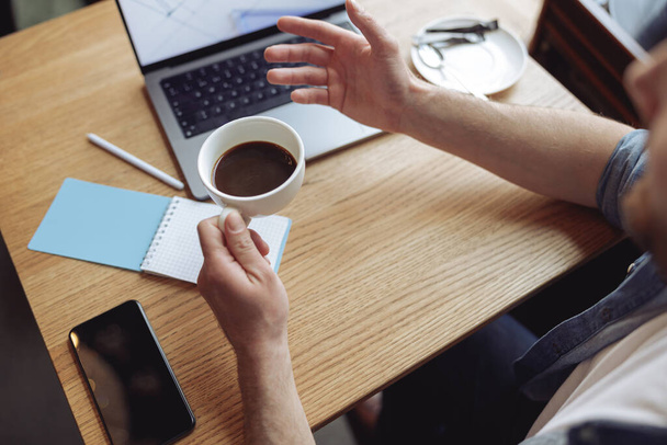 Close-up van witte kop zwarte koffie in mannelijke hand. Blanke man in cafe met laptop. - Foto, afbeelding