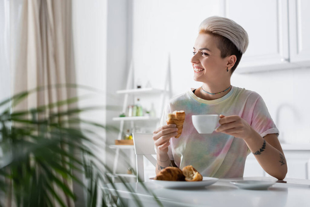 cheerful woman with coffee and delicious croissant looking away in kitchen - Photo, image