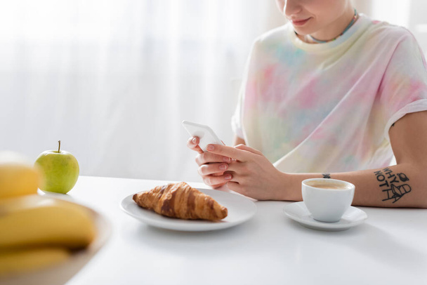 vista parcial de la mujer joven charlando en el teléfono inteligente cerca de café, croissant y manzana - Foto, imagen