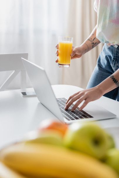 partial view of tattooed woman with orange juice typing on laptop near blurred fruits - Foto, Imagem