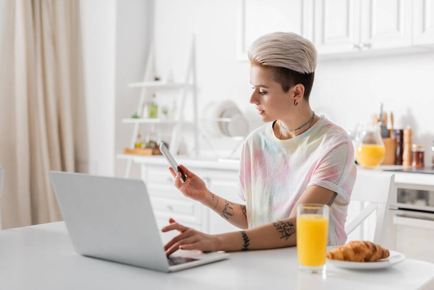 trendy tattooed woman with mobile phone typing on laptop near orange juice and croissant - Photo, image