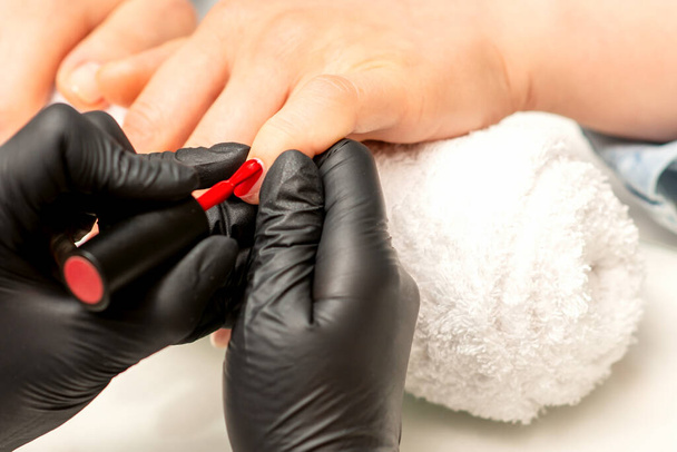 Фарбування манікюрним лаком. Крупним планом майстер манікюру в гумових чорних рукавичках наносить червоний лак на жіночий ніготь в салоні краси
 - Фото, зображення