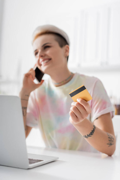 cheerful woman with credit card talking on mobile phone near laptop, blurred background - Foto, Imagen